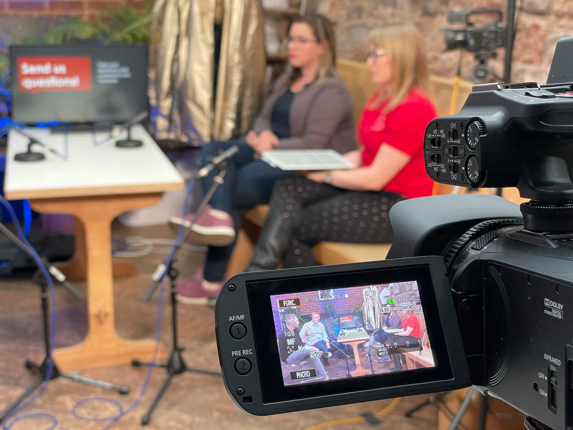 picture of a video camera (foreground right) with presenters in the background