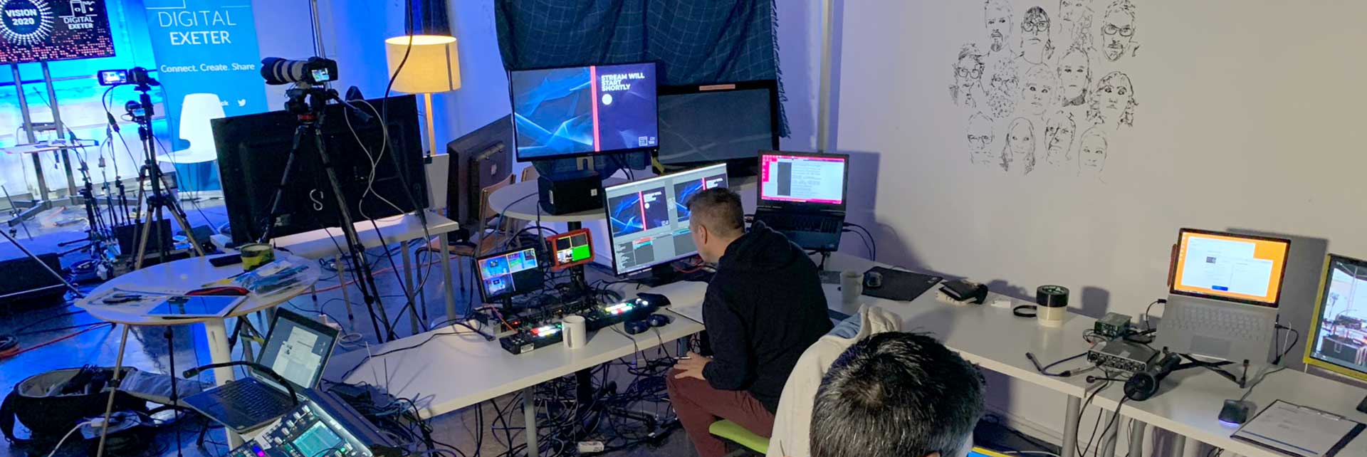 Behind the scenes photograph of computer screens, audio and video mixers and lots of cables